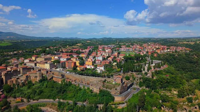 Establishing aerial shot of Pitigliano resting atop massive rock cliffs, surrounded by dense green vegetation and picturesque Italian hills under a bright cloudless sky.