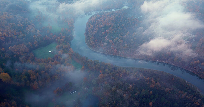 Aerial view of a winding river flowing through a dense forest with autumn foliage, partially obscured by low-hanging mist and clouds in Latvija.