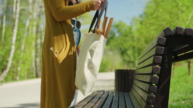 Woman in yellow cardigan approaches empty bench with art supplies. Woman places her bag on bench then sits down to prepare for painting outdoors outdoor