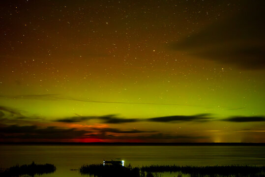 A spectacular night sky with the aurora borealis over a calm lake, clouds shimmering in greenish and golden light, and a dark shoreline with a small wooden pier.
