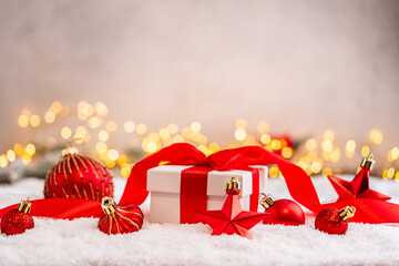Fototapeta premium A Christmas background featuring a white gift box tied with a red satin bow surrounded by red star and bauble ornaments on snow, softly lit by warm twinkling lights with copy space.
