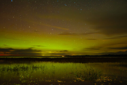 A spectacular night sky with the aurora borealis over a calm lake, clouds shimmering in greenish and golden light, and a dark shoreline with a small wooden pier.
