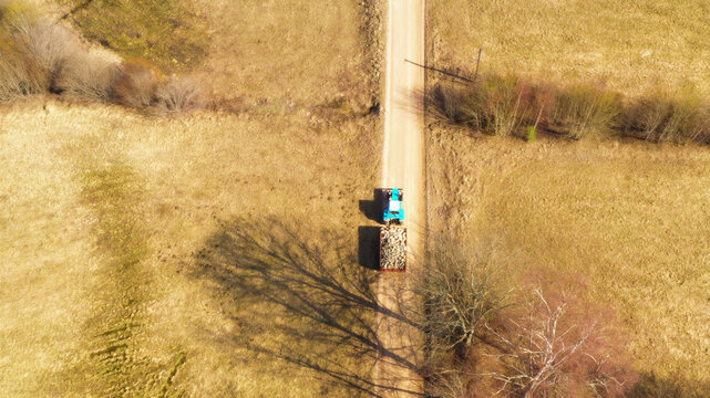 Aerial view of a blue tractor pulling a trailer loaded with logs on a dirt road through a rural landscape in Latvia.