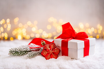 Fototapeta premium A Christmas background featuring a white gift box tied with a large red satin bow beside a frosted pine branch and red ornament on snow, softly lit by warm twinkling lights with copy space.