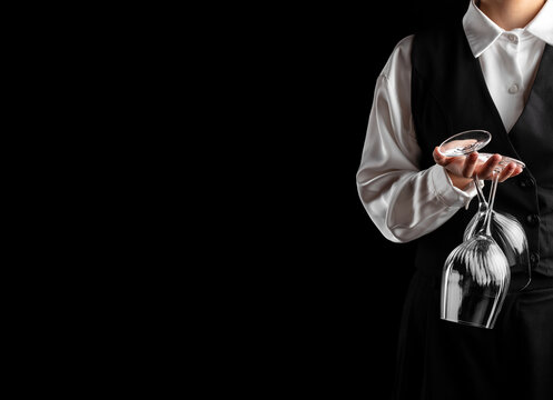 Waiter in white shirt and black vest holding a cocktail glass and inverted wine glass against a black background, cropped hospitality service portrait