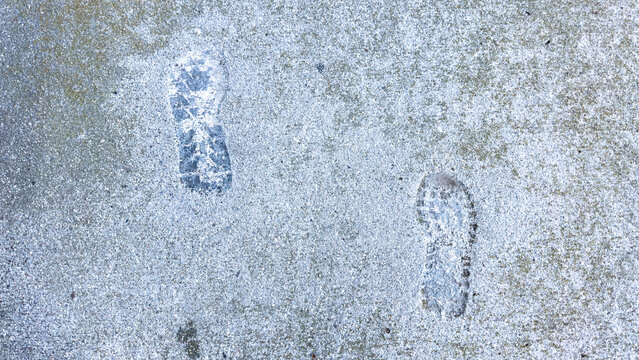 two wet shoe tread footprints mark a light grey gritty concrete sidewalk pavement showing distinctive sole impressions from footsteps across an outdoor hardscape walkway ground