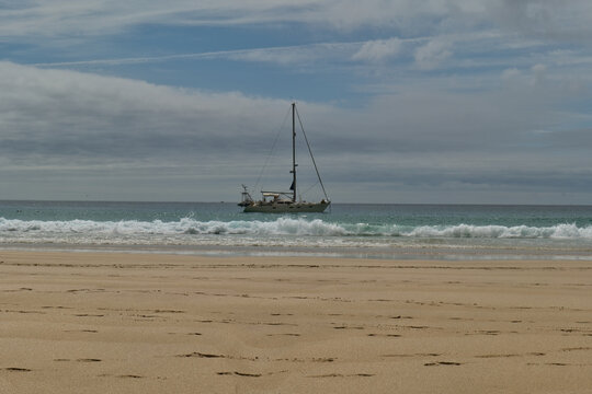 A sailboat is anchored close to a sandy beach with gentle waves breaking near the shore. The sky above is partly cloudy vb