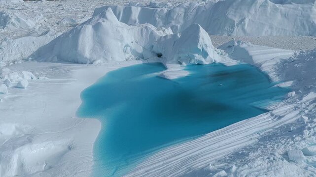 Aerial view of Ilulissat Icefjord featuring a vibrant turquoise meltwater pool surrounded by massive white icebergs under bright daylight in Ilulissat, Avannaata, Greenland.