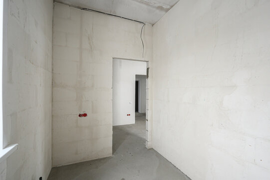 Sparse hallway in white-box finish shows exposed concrete block walls, raw doorways, visible wiring, and unfinished concrete floor with minimal fixtures