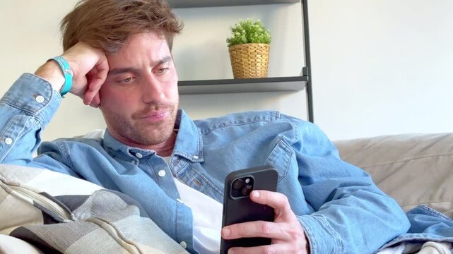 Young caucasian man sitting on the sofa, feeling bored and unmotivated while endlessly scrolling through social media feeds on his smartphone, procrastinating and wasting time at home