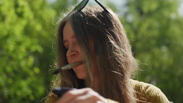 Close up view shows woman with shoulder length wavy hair wearing dark glasses resting atop their head. They hold various objects including what appears to be paintbrushes set against outdoor