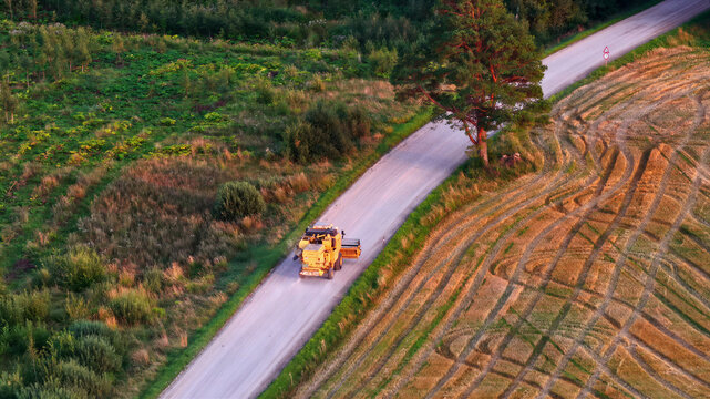 Aerial view of a combine harvester driving on a rural road next to a harvested field with tire tracks, surrounded by trees and greenery in Latvija.