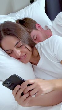 Young man waking up and suspiciously spying on his girlfriend's phone while she is smiling and texting, leading to an argument about jealousy and lack of trust in their relationship