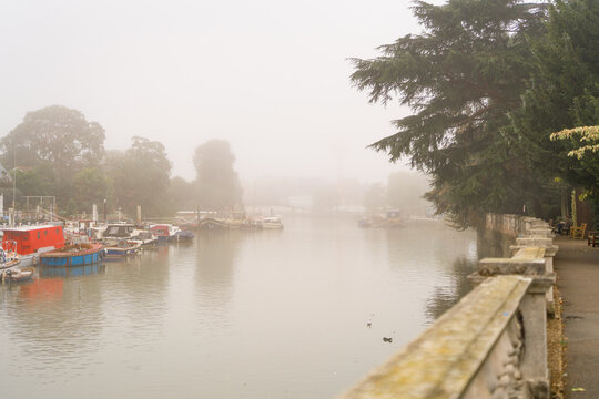 Atmospheric view of the River Thames with moored boats, trees and riverside buildings disappearing into the fog in Twickenham, London.