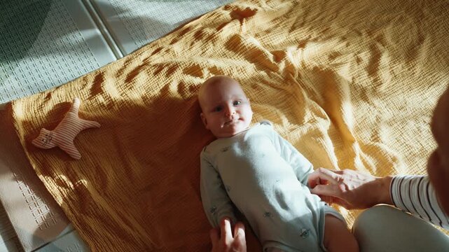 Young loving father doing developmental gymnastics with his cute newborn son on a yellow blanket in a sunlit room, supporting the infant's motor skills and early development