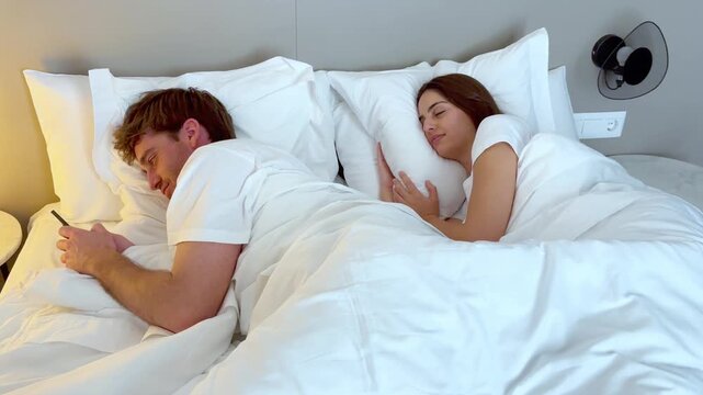 Distant young couple in bed at night, each separately engrossed in their smartphone, ignoring one another and illustrating modern relationship problems, phubbing, and technology addiction