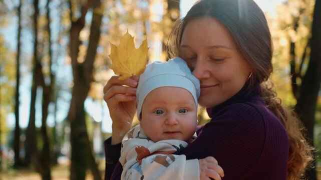 Young loving mother showing a yellow maple leaf to her curious infant son while holding him in her arms in a sunny autumn park, enjoying a beautiful day and creating lasting family memories