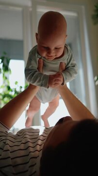 Loving father lifting his adorable newborn son in the air while lying on the floor. Cheerful baby smiling and laughing, enjoying a playful game with his dad in a sunlit room at home