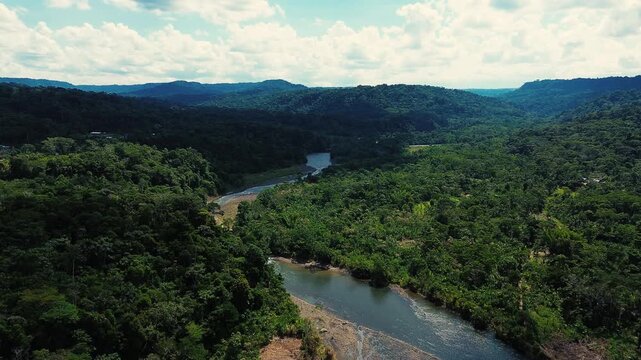 Drone footage looks across a winding river with exposed gravel and sediment bars, bordered by dense tropical rainforest in Ecuador's Oriente. Forested hills recede beneath a bright, cloud-filled sky, 