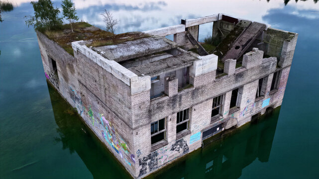 Aerial view of the submerged ruins of the Rummu quarry building with weathered brick walls, graffiti, and vegetation, surrounded by clear turquoise water in Rummu, Harju County, Estonia.
