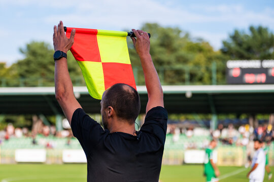Assistant referee holding up a red and yellow checkered flag high with both hands signaling substitution during an soccer match.