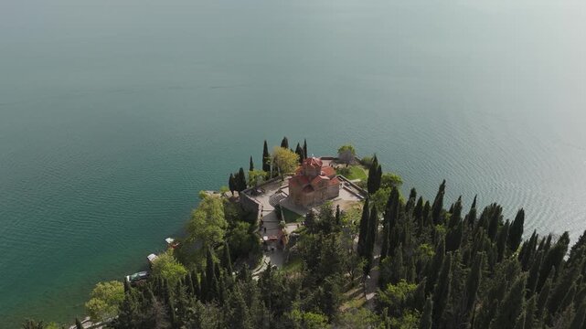 Sveti Jovan Kaneo, a Byzantine Orthodox landmark on a wooded cape in Ohrid, North Macedonia, surrounded by cypress trees, turquoise water, moored boats and footpaths