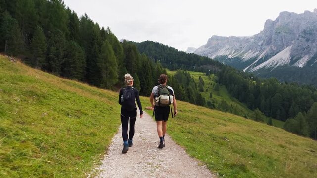 Walking follow shot behind two hikers with backpacks on meadow path toward rocky peaks in soft daylight, Dolomites, South Tyrol, Alps, Italy