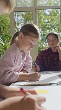 Vertical video of focused Caucasian girl drawing with pencil on paper during art class with diverse children. Footage suited for elementary education, creative workshop ads, afterschool programs
