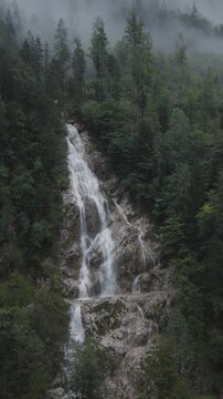 A vertical video aerial of the Plesnik waterfall with clouds and trees surrounding it, Logarska Dolina, Slovenia 