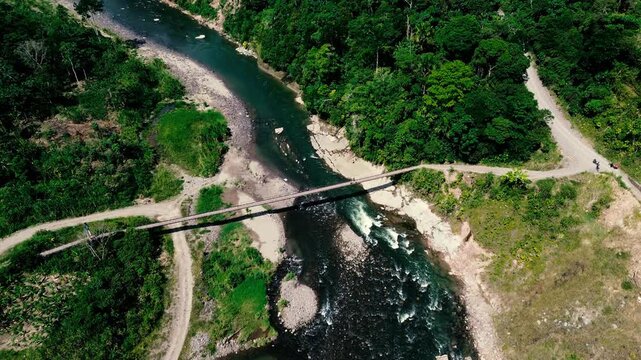 An overhead drone view shows a rocky river channel where green water breaks into white rapids among exposed stones and gravel bars. Dense tropical forest and narrow unpaved roads border the water in E