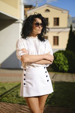 A woman with dark curly hair poses with crossed arms in a fashionable white outfit featuring a blouse and a high-waisted skirt with buttons