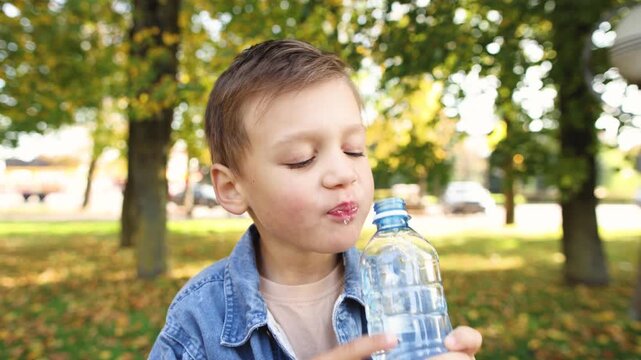 Young boy quenches thirst with water from plastic bottle in city park. Closeup shows refreshing drink after salty food and outdoor activity.