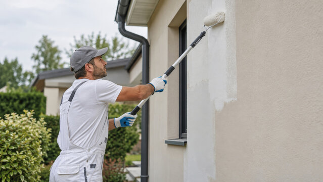 Painter applying fresh paint to an exterior house wall with an extension roller. Exterior painting and home maintenance. Renovation, repair and property upkeep. Painting contractor.