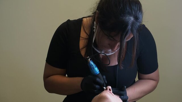 Eyebrow microblading shows a beauty technician applying pigment with a handheld precision device while wearing protective gloves and face protection, shown in slow motion.