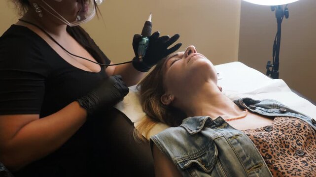 Preparing for eyebrow tattooing shows a beauty technician holding a cosmetic tattoo machine while positioning a client for a brow pigmentation procedure.