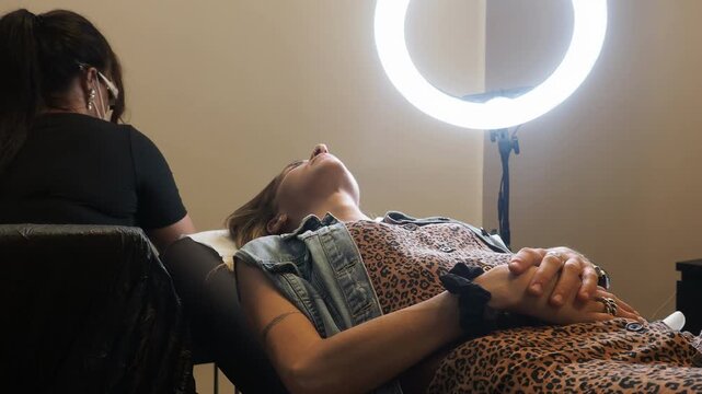 Eyebrow tattoo client reclines on a treatment bed while a beauty technician works beside her, with a ring light illuminating the cosmetic pigmentation procedure.