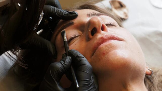 Eyebrow mapping shows a beauty technician defining the brow shape with a precision pencil while stabilizing the client's skin with gloved hands during cosmetic tattoo preparation.