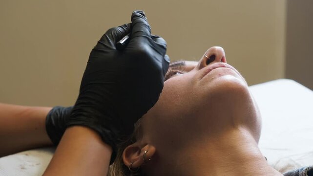 Eyebrow mapping shows a beauty technician holding a precision marking tool while stabilizing client's brow area with black gloves during cosmetic tattoo preparation. Eyebrow mapping, Brow shaping,