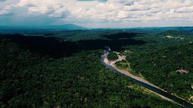 Drone footage gives an aerial view of a winding river bordered by dense tropical rainforest, with forested hills, gravel banks, scattered clearings and a few buildings in the valley. The landscape is 