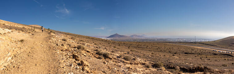 Panoramic view of the volcanic landscape around Montaña Blanca on Lanzarote, with a tourist walking in the background amidst arid terrain, volcanic mountains, and distant settlements. © Manoli Pérez
