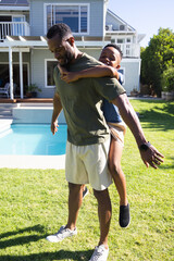 Naklejka premium African American father and school-age son playing piggyback on lawn by pool with smartwatch