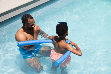 Naklejka premium African American father and son splashing in outdoor pool with pool noodles and sunglasses