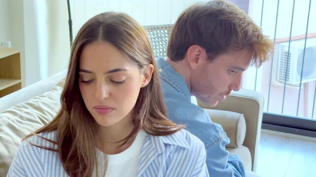 Stressed young couple sitting back to back on the sofa and arguing about their relationship problems and difficulties, representing a moment of crisis, frustration, and misunderstanding