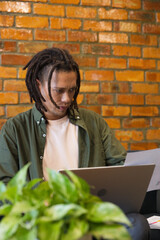 Dreadlocked man sitting at cafe table in green shirt holding paper, glancing at laptop beside plant © WavebreakMediaMicro