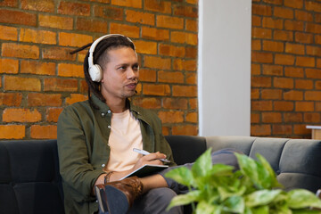 Man wearing white headphones sitting in lounge writing with pen in notebook near brick wall © WavebreakMediaMicro