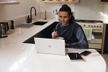 Man sitting eating from bowl while using white laptop at kitchen peninsula with notebook phone © WavebreakMediaMicro