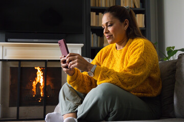 Fototapeta premium Mature African American woman sitting on sofa in living room by fireplace holding pink smartphone