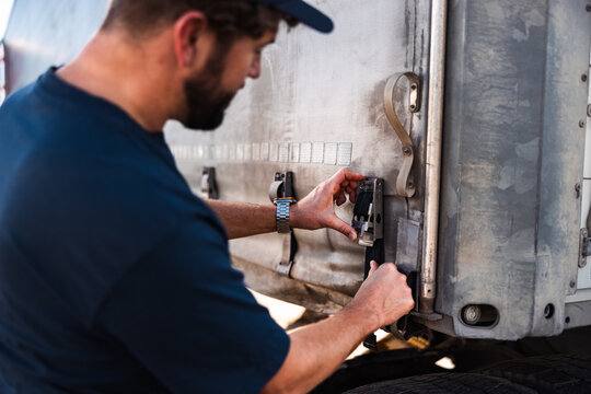 In a well-lit outdoor setting, a man focuses on fastening the latch of a truck trailer. The warm atmosphere highlights his concentration as he works diligently amid the vehicle's rugged exterior.