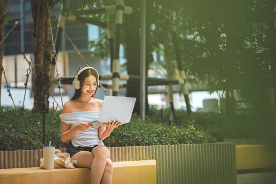 Young Asian woman using laptop outdoors while listening with headphones, sitting in green urban space with reusable tote bag and water bottle, enjoying remote work, digital lifestyle
