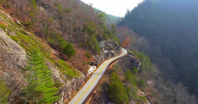 Scenic mountain road in North Carolina Highlands region. Car navigates curves beneath golden trees and steep rocky slopes in fall season.